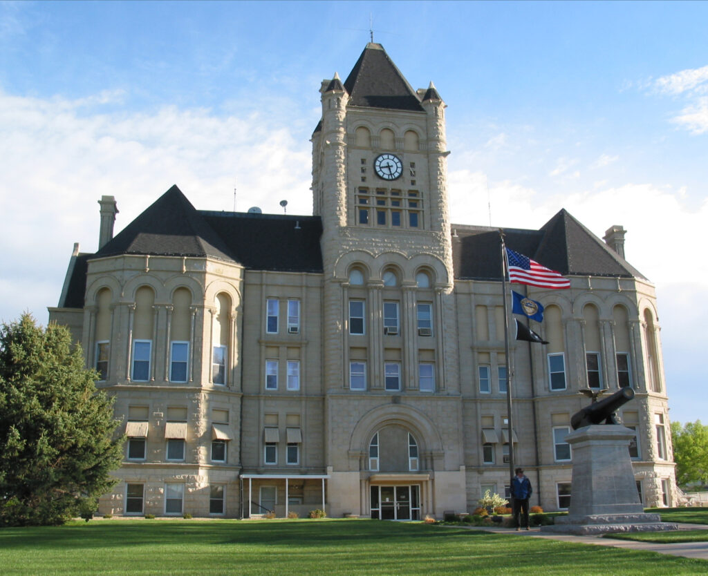 A low-angle view of the Gage County Courthoue in a grass field with an American flag.