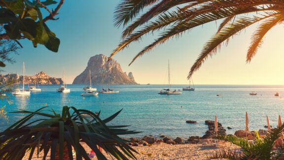 A view of the beach and boats on the water through palm trees.