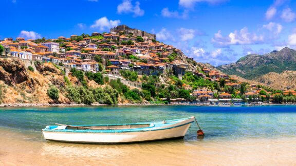 A small white and blue fishing boat sits in shallow water with the town of Mithymna in the background.