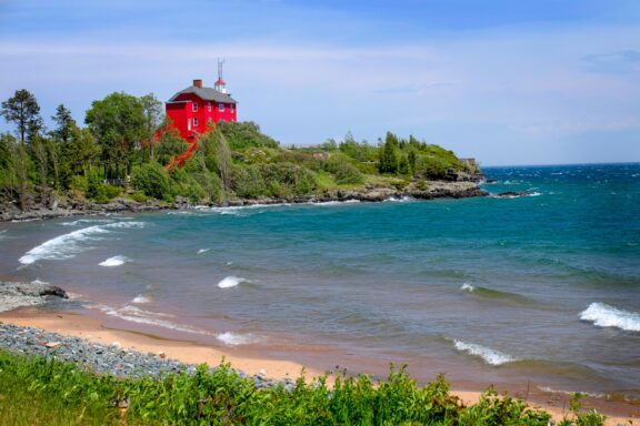 A large, red lighthouse sits atop a hill overlooking Lake Superior in Michigan.