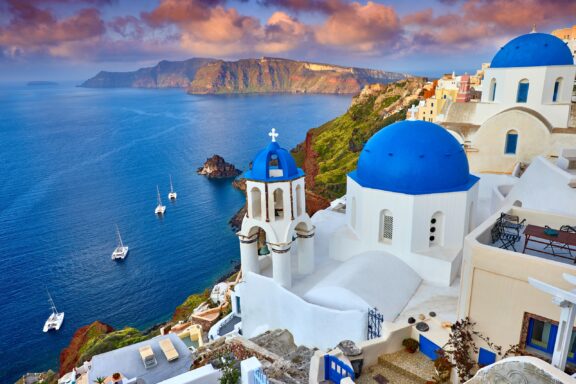 A view of white buildings with blue domes overlooking the water and distant cliffs from Greece’s island of Santorini.