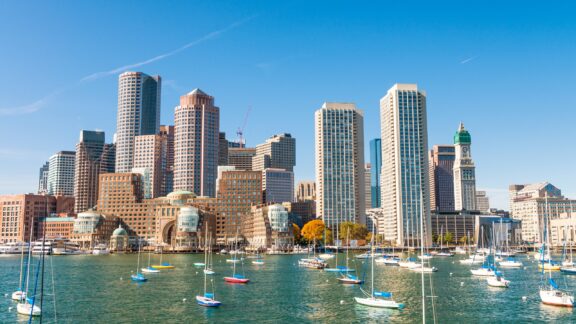 A low-angle view of the Boston skyline with water and boats in the foreground.