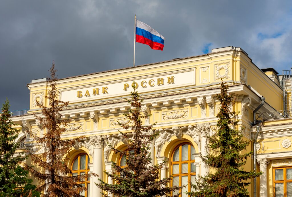 Russian flag waving above the Bank of Russia building with cloudy sky in the background.