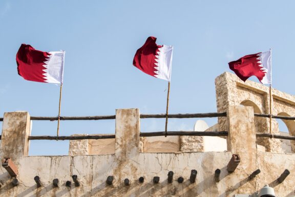 Three Qatari flags waving against a clear sky above a traditional stone wall.