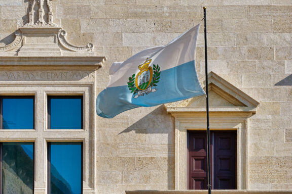 Flag of San Marino waving in front of a historical building with clear blue sky in the background.