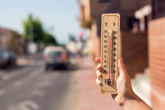 A hand holds a thermometer with a high temperature reading in a city. 