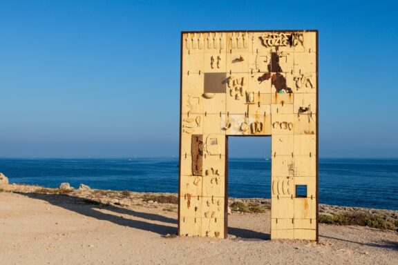 A tall sculpture of a doorway stands on the island of Lampedusa with the sea behind it.