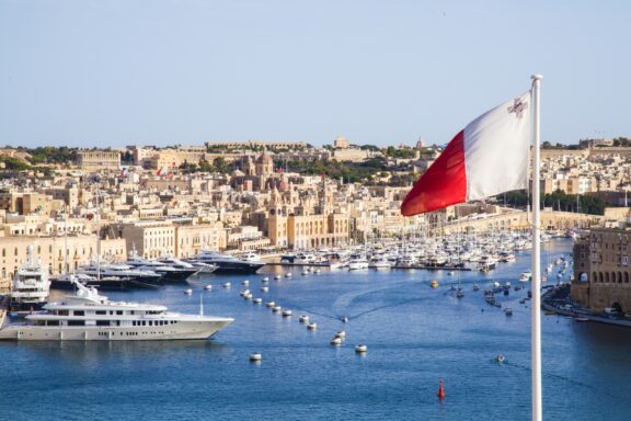 Maltese flag waving in the foreground with a view of a harbor and cityscape in the background.