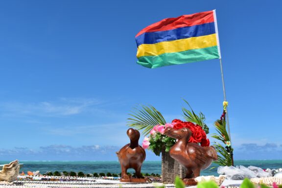 Flag of Mauritius waving against a clear blue sky with two wooden bird sculptures and tropical flowers in the foreground.