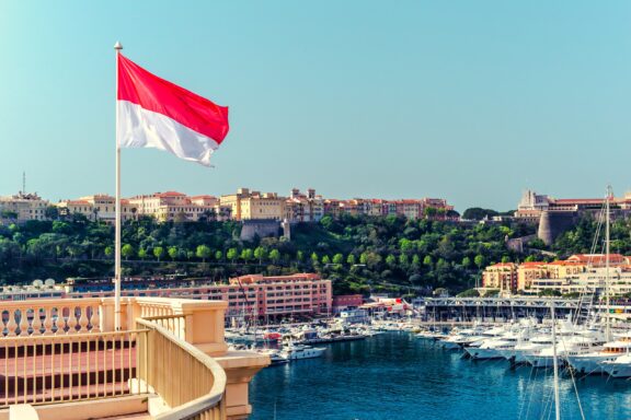 Flag of Monaco waving above a scenic view of the harbor with boats and buildings in the background.