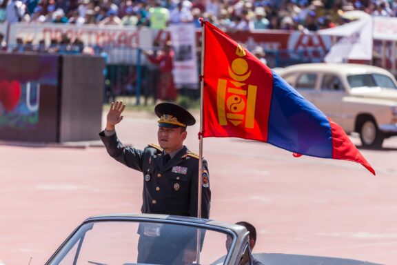 A uniformed officer saluting from a vehicle with the Mongolian flag waving in the foreground at a crowded event.