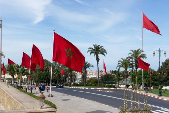 Red Moroccan flags with the national emblem fluttering above a city boulevard lined with palm trees under a clear blue sky.