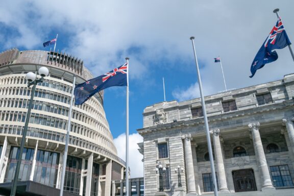 New Zealand flags flying in front of the Beehive and another government building in Wellington under a partly cloudy sky.