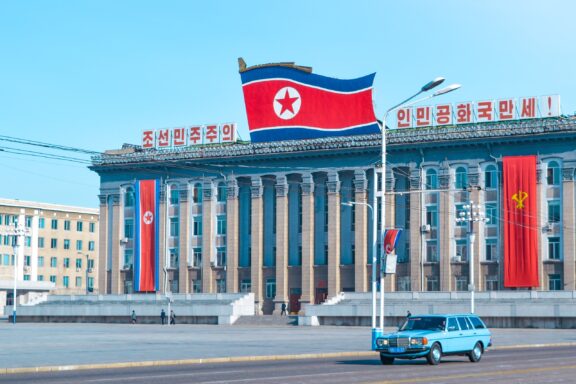 North Korean flag waving in front of a building with Korean script in Pyongyang, with a blue car passing by.