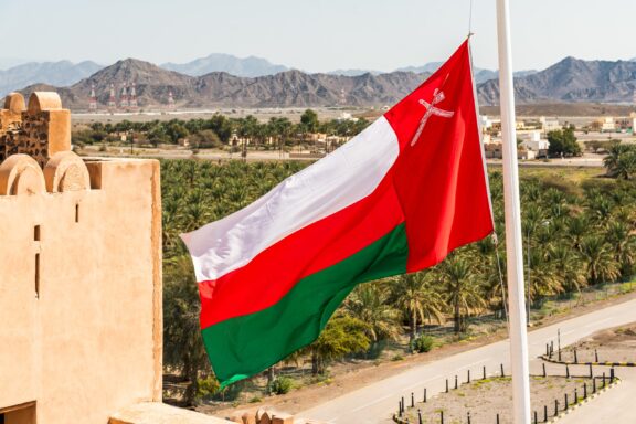 Oman flag waving with Jabreen Castle and mountains in the background.