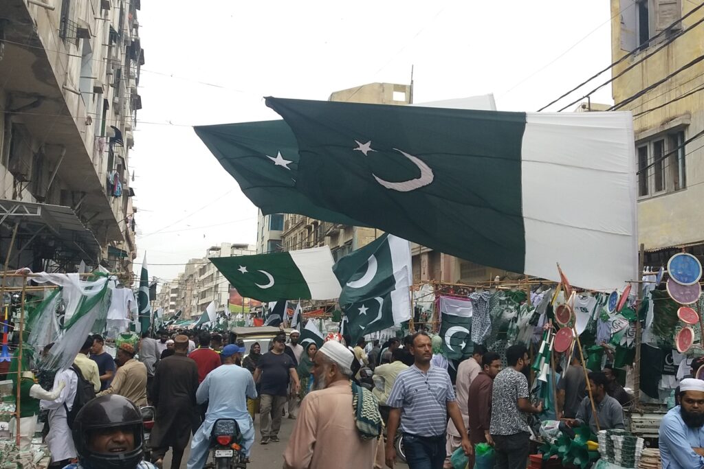 Pakistani flags being waved and displayed in a crowded street during Independence Day celebrations.