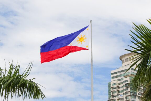 Philippine flag waving against a cloudy sky with palm fronds in the foreground and a building in the background.