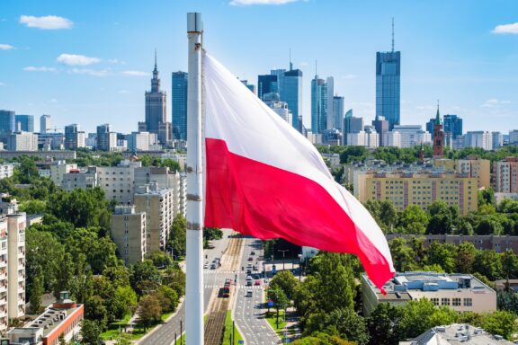 Polish flag waving in the foreground with the skyline of Warsaw, including modern skyscrapers and greenery, in the background.