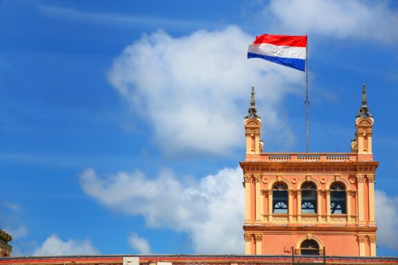 Flag of Paraguay waving above the pink facade of the Presidential Palace against a blue sky with clouds.