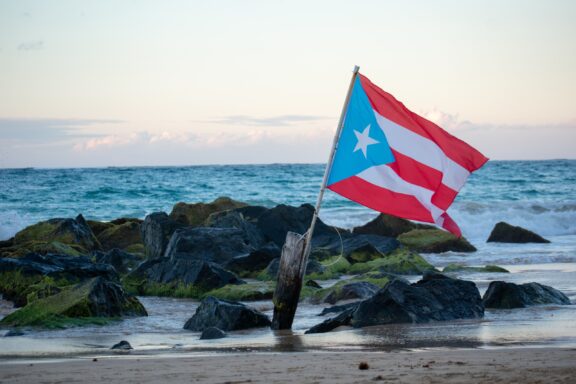 Puerto Rican flag waving on a pole among rocks at the beach with the ocean in the background.