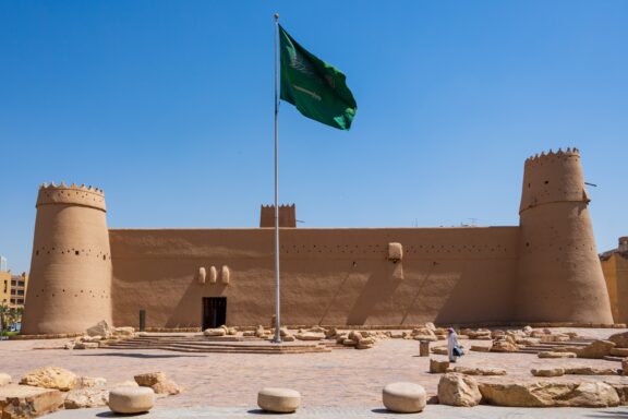 Saudi Arabian flag waving above an ancient fort under a clear blue sky.