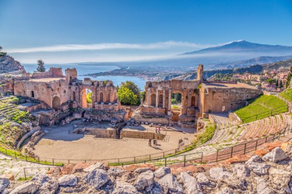 Mount Etna can be seen in the distance, and the ruins of a stone theater occupy the foreground.