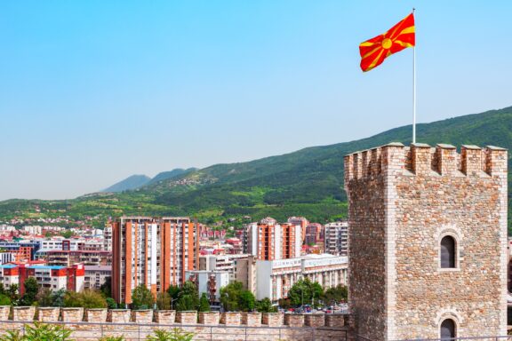 Macedonian flag waving above Skopje Kale Fortress with cityscape and mountains in the background.