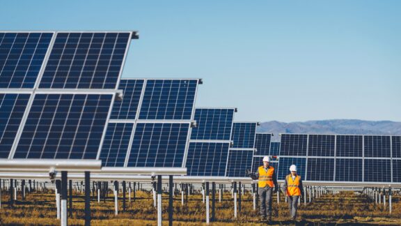 Two workers in orange vests walk next to solar panels in a solar power station.