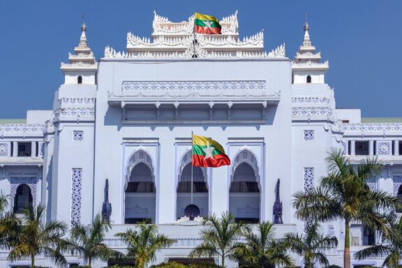 White colonial-style building with a tiered roof and a Myanmar flag flying in front.