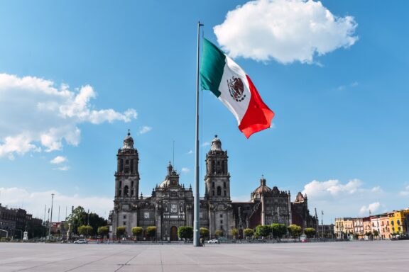 Mexican flag waving in front of the Metropolitan Cathedral at Zócalo Square, Mexico City, under a clear blue sky.