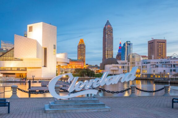 A white sign that reads, “Cleveland” stands next to the water with the Cleveland skyline in the background.