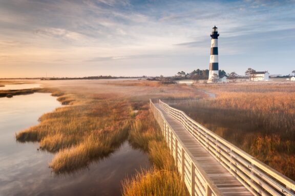 A wooden boardwalk travels through a marsh toward Bodie Island Lighthouse.