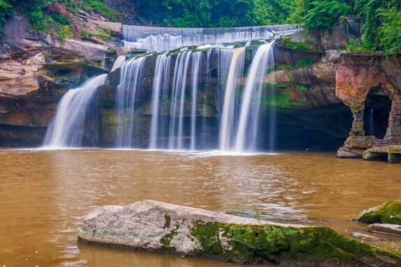 Water flows over small cliffs surrounded by green plants.