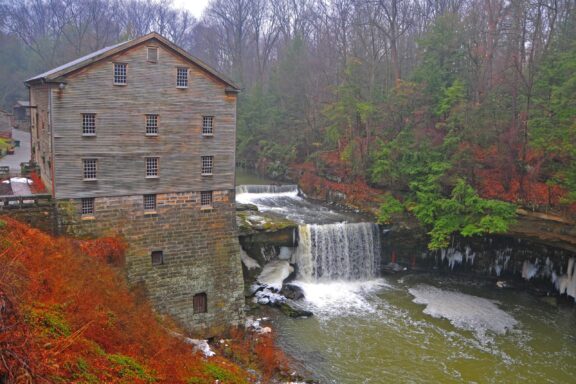 A large brick and wood mill stands next to a small river on a winter day.