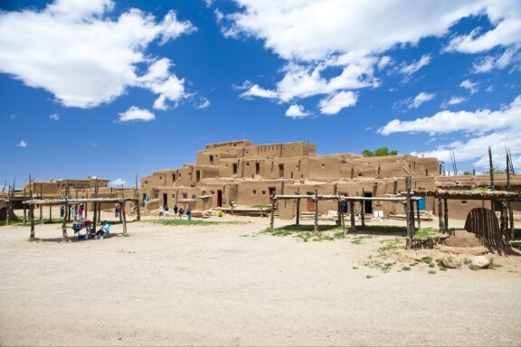 Several adobe buildings stand together in Taos County under a partly cloudy sky.