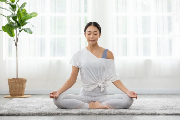 A woman sits in meditation on a carpet in a white room.