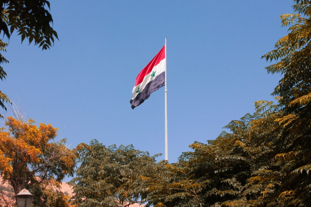 The Syrian flag waving against a clear blue sky with trees in the background.