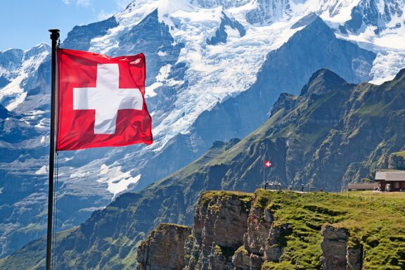 Swiss flag waving in front of snow-capped mountains and green hillsides.