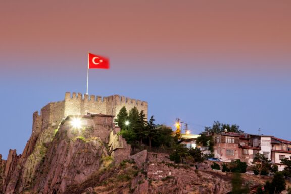 Turkish flag waving above an ancient fortress at dusk with illuminated walls and surrounding buildings.