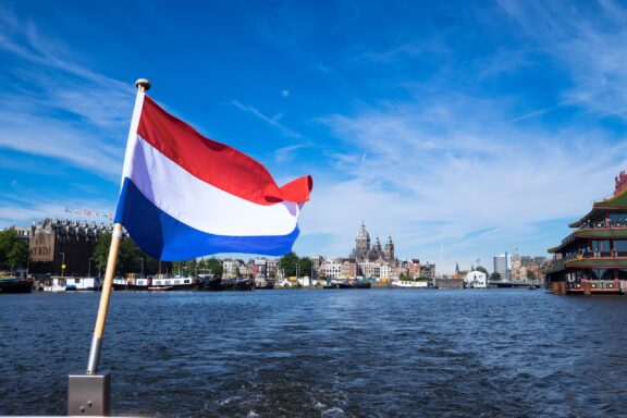 Netherlands flag waving in front of a canal with Amsterdam architecture in the background.