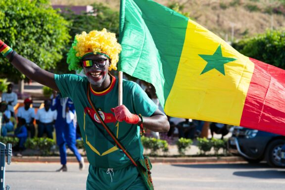 A person wearing a yellow wig and sunglasses, holding the Senegal flag, and dressed in green and red sports attire, celebrates enthusiastically on a sunny day.