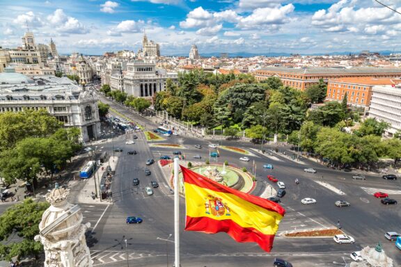 Aerial view of a bustling city intersection with the Spanish flag prominently displayed in the foreground.