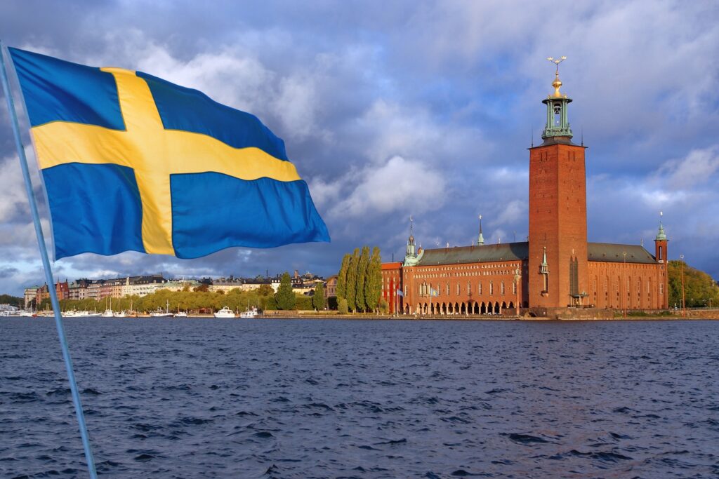 Swedish flag waving in the foreground with the Stockholm City Hall in the background under a cloudy sky.