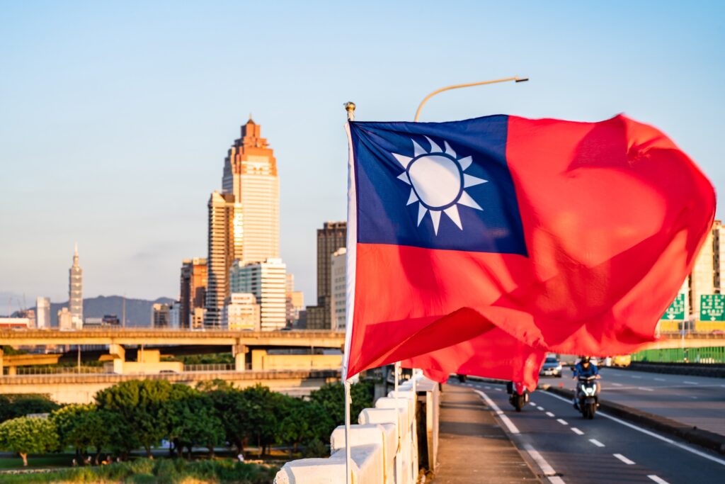 Taiwanese flag waving in the foreground with Taipei city skyline and clear blue sky in the background.