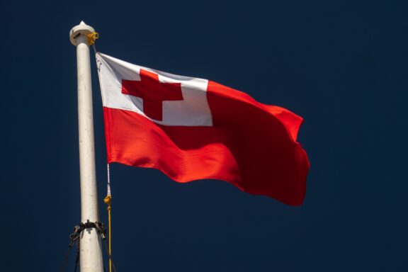 The flag of Tonga waving on a flagpole against a clear blue sky.