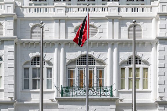 Flag of Trinidad and Tobago waving in front of a white colonial-style building with arched windows and decorative railings.