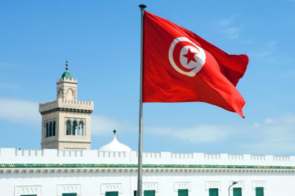 Tunisian flag waving in front of a building with a clock tower under a clear blue sky.