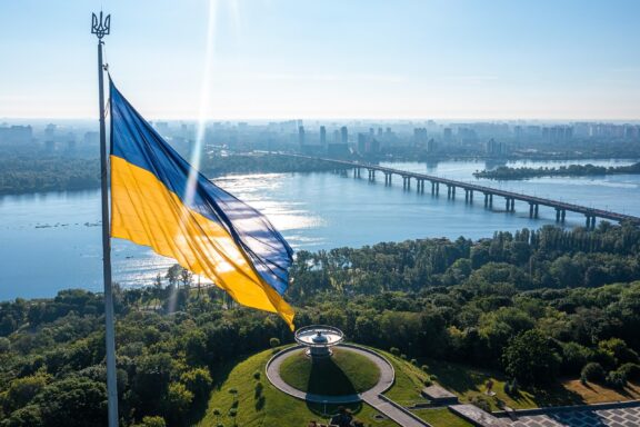 Ukrainian flag waving in the foreground with a panoramic view of Kyiv and a river in the background.