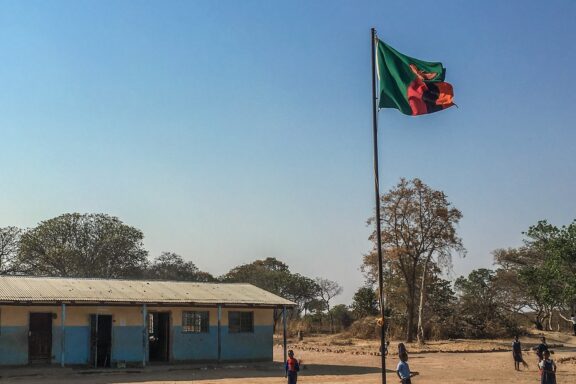 Zambian flag waving on a pole with a clear sky, in front of a school building with children playing outside.