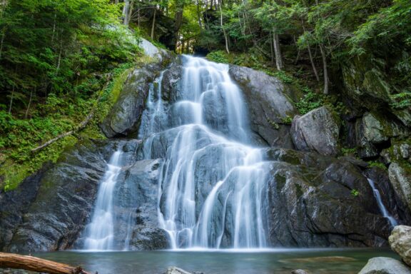 A serene waterfall cascades over smooth, mossy rocks surrounded by lush greenery.